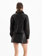 Chaqueta de cuero ovino color negro, con textura graneada, silueta de hombros caídos y largo a la cadera. Presenta recortes en el delantero y la espalda, dos bolsillos superiores con tapa y dos bolsillos laterales, cierre frontal con botones metálicos y cuello camisero.