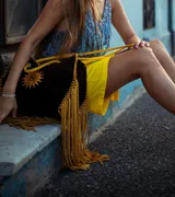 Bolso tipo tote de gamuza color negro con flecos y bordes en color salmón. Presenta un estampado de un sol con las iniciales de la marca en el centro. Tiene correa larga del mismo material.