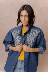 Camisa de denim azul con bordados florales en tonos amarillos y celestes sobre los hombros y el pecho. Presenta cuello clásico, cierre frontal con botones y mangas largas con puños.