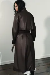 Gabardina larga de efecto encerado en color marrón oscuro, con cuello solapa y cierre frontal cruzado con botones. Presenta mangas largas con puños vueltos en pana, bolsillos laterales y cinturón ajustable de pana. Incluye pinza en la espalda.