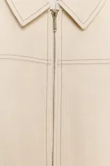 Chaqueta crop color beige con cuello solapa, manga larga y cierre frontal con cremallera. Presenta detalles de costuras a contraste en color marrón claro.