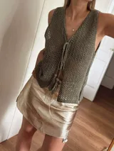 Chaleco tejido en punto calado con hilo de algodón y lurex, de color negro con destellos plateados. Presenta escote profundo en V, cierre frontal con cordones entrelazados terminados en borlas y un corte recto y holgado.