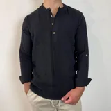 Camisa de manga larga confeccionada en muselina de algodón, color negro. Presenta cuello mao y cierre frontal parcial con tres botones de madera. Las mangas incluyen una trabilla lateral para sujetar el puño cuando se lleva arremangada.