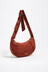Bolso tipo hobo de la marca Bimba y Lola, modelo Moon mediano, de color rojo óxido. Confeccionado en nylon con diseño de media luna, cierre superior con cremallera, correa ajustable con hebilla metálica y el logotipo de la marca impreso en la parte frontal.