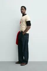 Camiseta de corte regular, color crema, con cuello redondo y ribetes en las mangas y cuello en color negro. Presenta un estampado frontal con un corazón rojo y texto en negro que dice "Everything beats for something".