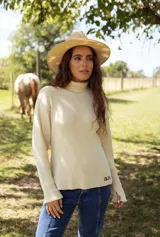 Polera de tejido de punto acanalado color beige, con cuello alto, manga larga y detalle de logo bordado en el ruedo.