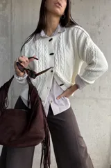Cardigan de punto trenzado color natural, con cuello redondo, cierre frontal de botones negros y mangas largas con puños acanalados.