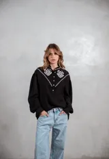 Camisa negra de manga larga con bordados blancos en la parte superior delantera y hombros. Presenta cuello mao, cierre frontal con botones y mangas con puños ajustados.