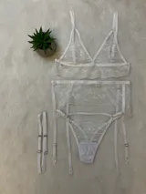 Conjunto de lencería de encaje blanco compuesto por un bralette triangular, una tanga con tiras laterales y un portaligas integrado. El diseño presenta detalles de encaje floral y breteles regulables.