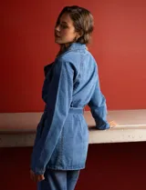 Chaqueta larga de jean azul con cuello con solapa, abertura central con botones, bolsillos delanteros, cintura con pasadores y lazo de ajuste, y puños con botón.