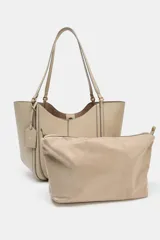 Bolso tipo shopper de hombro en color beige, con diseño estructurado, doble asa y detalle de pespuntes verticales en el cuerpo. Incluye un colgante decorativo tipo etiqueta y cierre imantado.