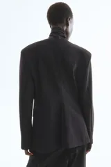Blazer de punto grueso negro, con escote en V profundo sin cuello, hombreras y cintura entallada. Presenta cierre de un botón frontal y bolsillos delanteros con solapa.