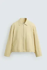 Cazadora de tejido con algodón, color amarillo pálido, con cuello solapa y cierre frontal de cremallera. Presenta bolsillos frontales de vivo y bajo ajustable con elástico en los laterales.