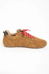 Zapatilla de estilo deportivo color camel, confeccionada en cuero gamuzado. Presenta cordones rojos con detalles blancos y un pequeño refuerzo gris en el talón.