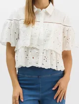 Camisa de algodón blanca con cuello clásico y cierre frontal de botones. Presenta un diseño con capas superpuestas y volados, combinando paneles de tela lisa con tejido bordado suizo de motivos calados y una cenefa decorativa. Posee mangas cortas y una silueta acampanada.