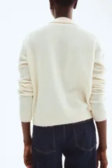 Cárdigan corto de punto suave color crema, con cuello camisero, abotonadura frontal con botones marrones y remate acanalado en puños y bajo.