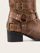 Bota de caña alta estilo biker, color marrón, con punta redondeada y taco cuadrado. Presenta múltiples tiras horizontales con hebillas metálicas y detalles de herrajes en el lateral. Confeccionada en cuero con forro vacuno y suela de madera.