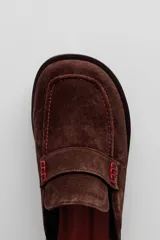 Mule tipo mocasín de gamuza en color tostado, con detalle de costuras decorativas y suela plana de goma negra.