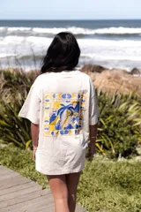 Remera oversize color crudo con estampado en la espalda de un paisaje tropical con palmeras, sol y mar en tonos amarillo, azul y celeste.