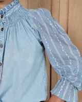 Camisa de denim celeste con detalles de broderie en las mangas, cuello alto con volados y canesú con frunces. Presenta cierre frontal con botones y mangas largas con puños elásticos.