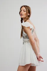 Enterito corto blanco de tela liviana, con escote en V, canesú de encaje con bordados brillantes, volados en los tirantes y diseño de short con falda con vuelo.