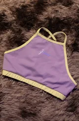 Top deportivo color lila con ribetes en amarillo claro. Presenta tirantes finos y un logo estampado en el centro en tono lila más oscuro.