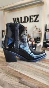 Botas tejanas de cuero charolado negro, con taco cuadrado de 7 cm y punta fina. Cuentan con cierre interno y costuras visibles en la suela.