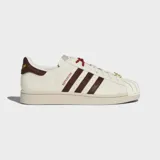 Championes Adidas Superstar II con exterior de cuero y material sintético en color blanco crema. Presentan las icónicas tres tiras laterales y refuerzo en el talón en color marrón oscuro texturizado, puntera de goma con relieve y suela de caucho al tono.