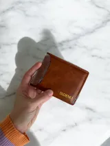 Billetera de eco cuero color marrón con tarjetero extraíble integrado. Presenta un diseño compacto con ranuras para tarjetas y logo grabado en tono dorado.