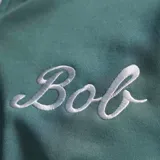 Campera deportiva Adidas con cierre frontal, cuello alto, mangas largas con las icónicas tres tiras blancas y puños elásticos. Presenta el logo de Adidas en el pecho derecho, la palabra 'Bob' bordada en el pecho izquierdo y un parche conmemorativo en la parte inferior derecha.