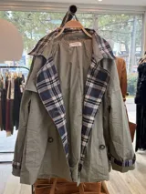 Gabardina de corte clásico en color verde militar, con solapas amplias y puños con detalles en estampado de cuadros escoceses en tonos azul, blanco y negro. Presenta cierre frontal cruzado con botones y bolsillos laterales.