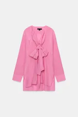 Blusa fluida de color rosado con cuello redondo ajustable mediante lazadas del mismo tejido, manga larga con puño y bajo asimétrico con aberturas laterales.