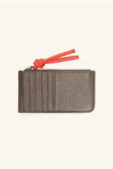 Monedero rectangular de cuero vacuno con textura granulada en color marrón topo. Cuenta con cierre superior metálico y un tirador decorativo de cuero en color naranja con un nudo. Presenta un pequeño logo grabado en el centro.