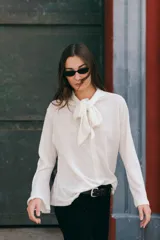 Blusa de lino color crudo, con cuello alto y lazo decorativo frontal. Presenta mangas largas con corte acampanado y diseño entallado con pinzas delanteras.