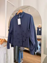 Camisa de algodón color azul marino, con cuello redondo, botonera oculta y apliques en relieve distribuidos en el frente. Presenta mangas largas con puños abotonados y corte recto.