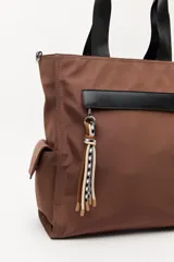 Bolso tipo tote color negro, con doble asa superior y correa desmontable. Presenta un bolsillo frontal con solapa, bolsillos laterales y un detalle colgante decorativo con tiras de colores.