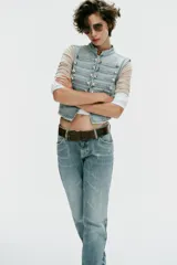 Chaleco de denim color celeste con cuello subido, sin mangas, con detalle de aplicaciones horizontales y botones metálicos en el frente. Cuenta con cierre frontal con ganchos metálicos ocultos.