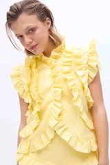 Blusa amarilla sin mangas, con cuello alto y cierre frontal abotonado. Presenta abundantes volados decorativos a lo largo de la abertura y en los hombros.