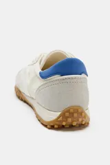 Zapatilla deportiva estilo blucher de color blanco, con detalles en gris claro, una franja lateral blanca y una talonera azul. Cuenta con cordones, punta redonda y suela de goma con tachuelas.