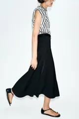 Vestido midi combinado con parte superior de camisa a rayas verticales blancas y negras, cuello solapa, manga sisa con frunces y botones dorados. La parte inferior es una falda midi negra de corte recto. Incluye un cinturón negro con textura de cocodrilo y hebilla dorada.