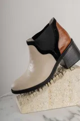 Botineta de diseño tricolor con cuerpo en tono beige, panel elástico lateral negro y talón con textura símil reptil en color marrón. Presenta un taco cuadrado robusto de color negro.