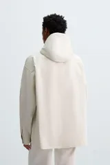 Sobrecamisa de corte oversized confeccionada en tejido compacto de algodón. Presenta cuello subido con capucha ajustable mediante cordones, cierre frontal con botonadura, manga larga con puños abotonados y dos bolsillos de plastrón en la parte frontal.
