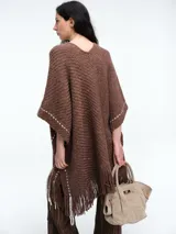 Poncho tejido de punto grueso color marrón, con escote en V, diseño amplio y flecos en el ruedo. Presenta detalles de costuras decorativas en los laterales.
