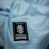 Camiseta de fútbol celeste con cuello en V y mangas cortas con ribete blanco. Escudo de Uruguay bordado en el pecho.