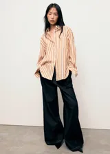 Camisa de corte oversize con diseño de rayas verticales en tonos blanco y naranja. Presenta cuello camisero clásico, cierre frontal con botones, bolsillo de parche en el pecho y mangas largas con puños abotonados.