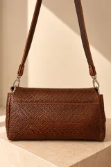 Cartera de cuero vacuno color marrón oscuro, con diseño trenzado y solapa frontal con placa metálica dorada grabada con la marca PECARÍ. Cuenta con correa larga para hombro y herrajes metálicos plateados.