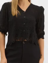 Blusa de algodón semitransparente con diseño floral bordado a tono. Presenta escote en V, cierre frontal con botones y mangas al codo ligeramente abullonadas. Incluye detalles de encaje calado estilo macramé en los puños y en el ruedo con terminaciones en ondas.