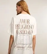 Remera oversized de algodón color crudo, con cuello redondo y mangas cortas. Estampa trasera con texto "Amor Peligroso" y listado de ciudades con fechas, en tipografía estilo western.
