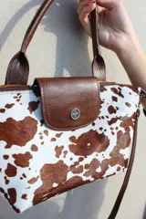 Cartera tipo bolso de mano confeccionada en cuero gamuzado color suela, con solapa de cuero liso y cierre metálico. Incluye doble manija corta y correa larga desmontable.