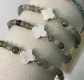 Pulsera elástica confeccionada con cuentas de piedra natural gris y un dije central en forma de trébol de nácar blanco.